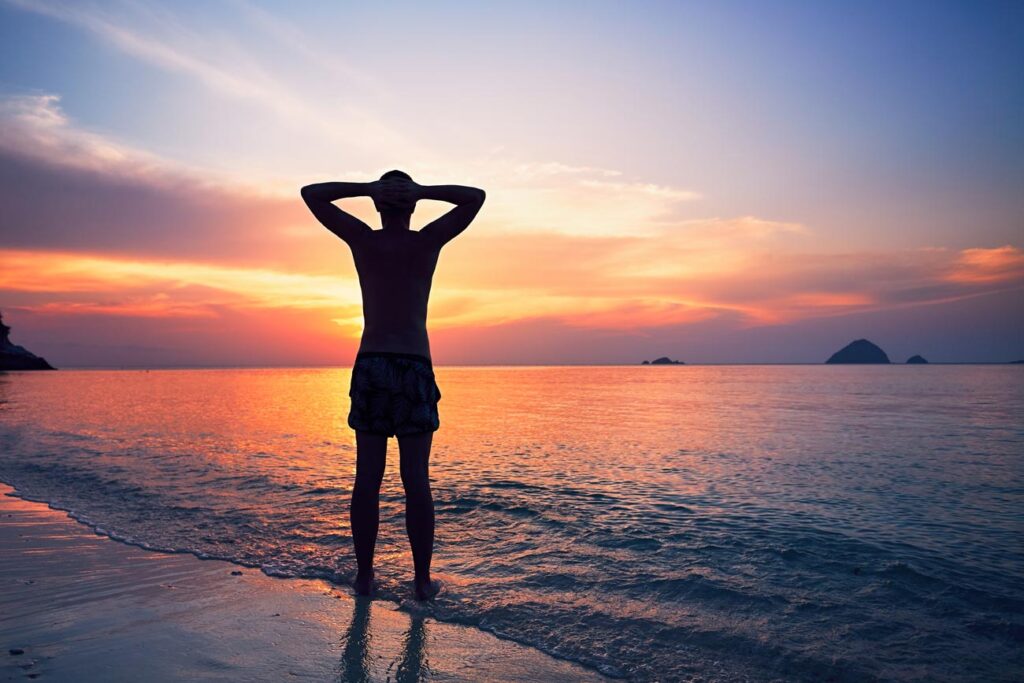 Alone on the beach. Pensive young man standing in water of the tropical sea during beautiful sunset.