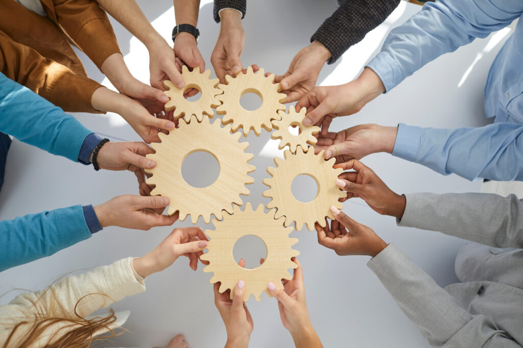 People holding wooden gears that symbolize well-coordinated teamwork. Top view close up of hands of people standing in circle.